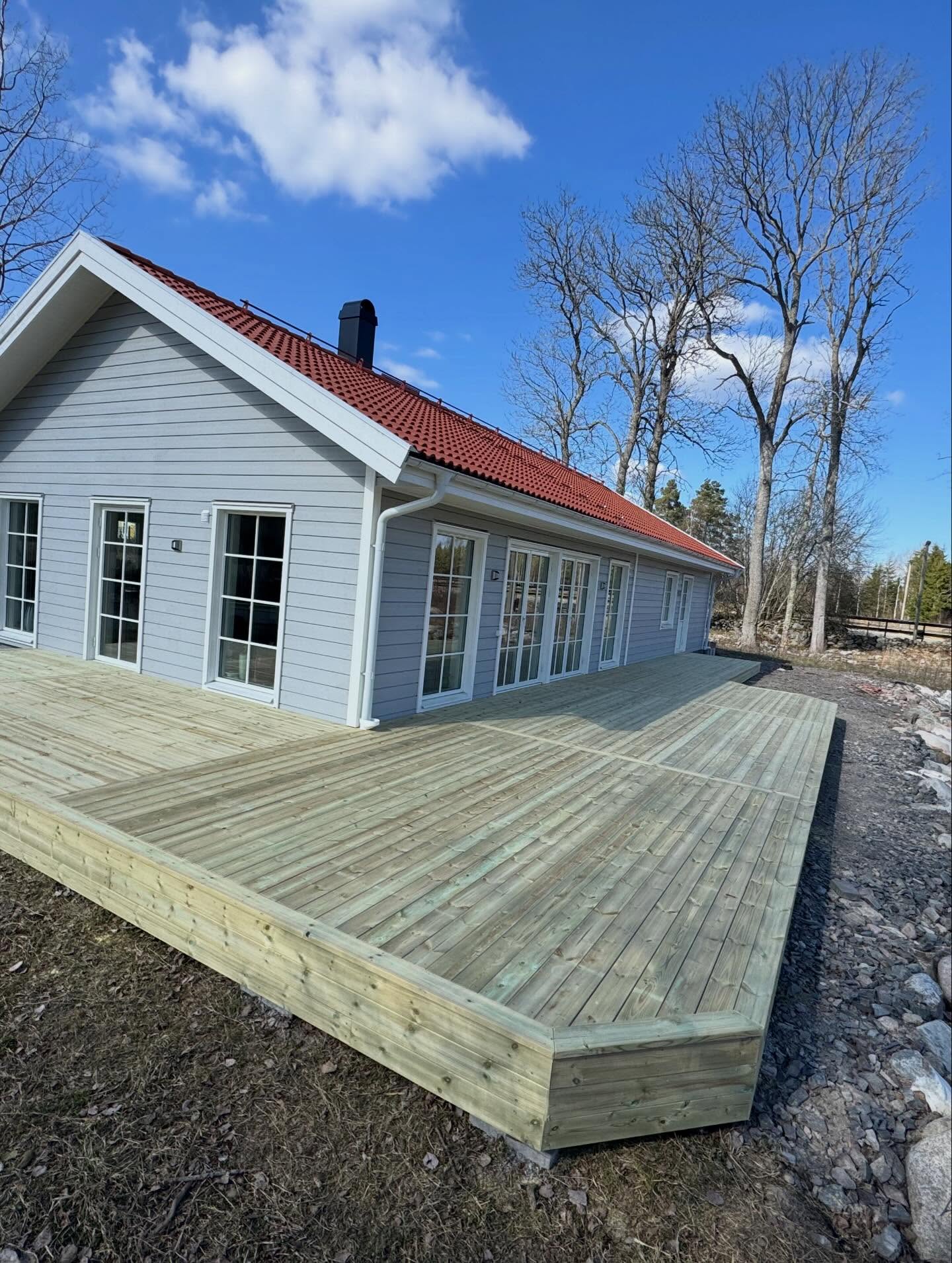 Omlöpande trädäck längs grå villa med vita pardörrar och rött tegel tak